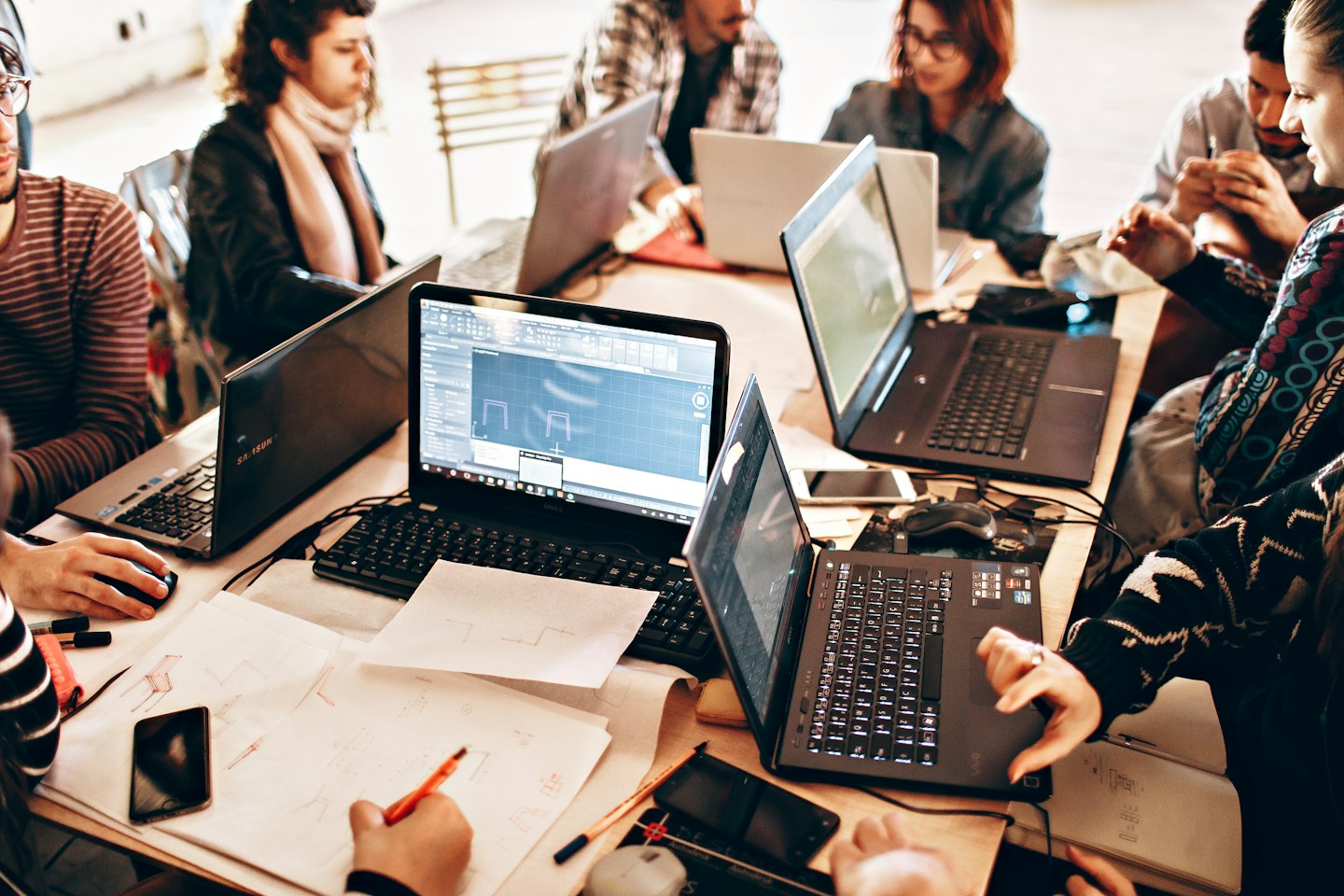 En grupp unga, mångkulturella vuxna som samarbetar vid en träbord med laptops.